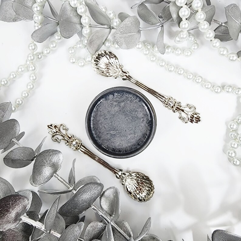 May include: A small, round, gray container with a black lid, surrounded by silver eucalyptus leaves and a pearl necklace. Two silver spoons with ornate handles are placed on either side of the container.