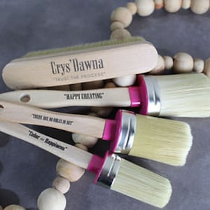 May include: Set of four paint brushes with wooden handles and pink ferrules. The handles are engraved with inspirational quotes, including "Happy Creating" and "Color = Happiness". A wooden brush cleaning tool is also included.