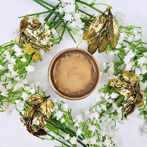 May include: A gold-toned metal coaster with a round, slightly raised edge. The coaster is surrounded by white flowers and gold-colored leaves.