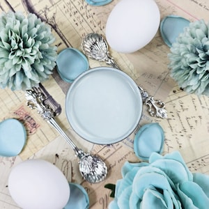 May include: A white ceramic bowl with a light blue glaze, surrounded by decorative elements including blue flowers, eggs, and silver spoons.