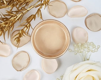 May include: A gold jar with a lid, filled with a light beige cream. The jar is surrounded by light beige silk flower petals and a sprig of white pearl-like beads.