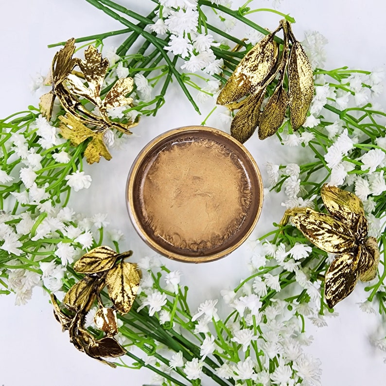 May include: A gold-toned metal coaster with a round, slightly raised edge. The coaster is surrounded by white flowers and gold leaf sprigs.
