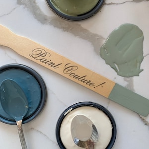 May include: Three paint samples in small jars with lids, a white sample, a blue sample, and a green sample. A wooden paint stir stick with the text "Paint Couture!" is lying on a white marble surface. A spoon is in each jar.