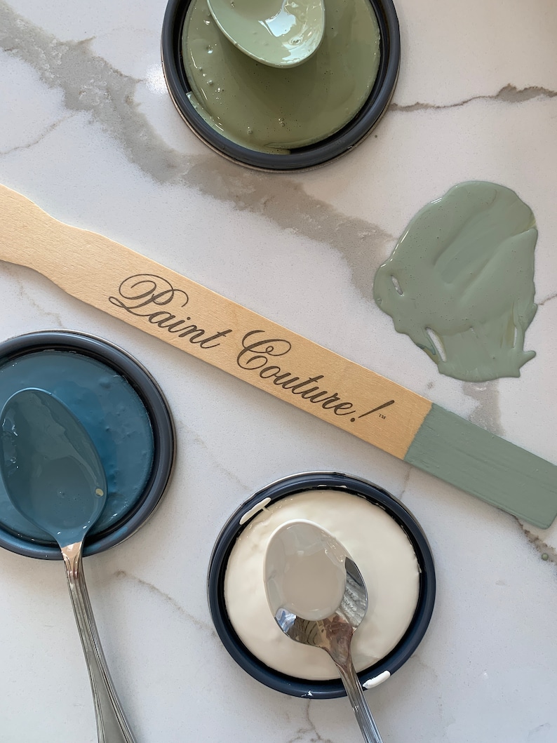 May include: Three paint samples in small jars with lids, a white sample, a blue sample, and a green sample. A wooden paint stir stick with the text "Paint Couture!" is lying on a white marble surface. A spoon is in each jar.