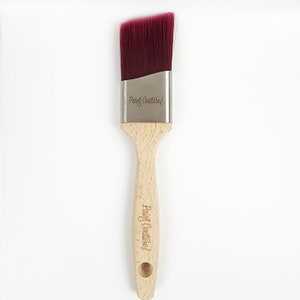 Pu&ograve; includere: Un pennello per dipingere con manico in legno e una testa di setole inclinata di colore bordeaux. Il pennello ha le parole "Paint Couture" incise sul manico e sulla virola metallica.