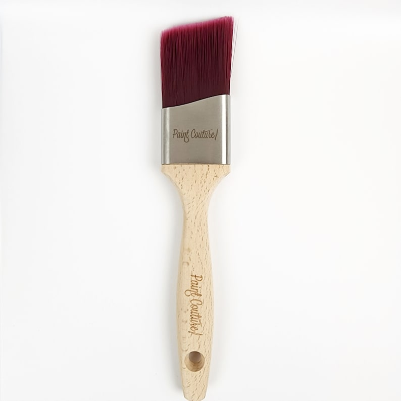 Pu&ograve; includere: Un pennello per dipingere con manico in legno e una testa di setole inclinata di colore bordeaux. Il pennello ha le parole "Paint Couture" incise sul manico e sulla virola metallica.