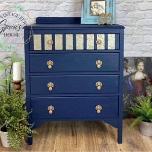 May include: A navy blue chest of drawers with four drawers and gold-toned hardware. The top drawer has a decorative gold and white patterned paper inlay. The chest of drawers is sitting on a wooden floor.