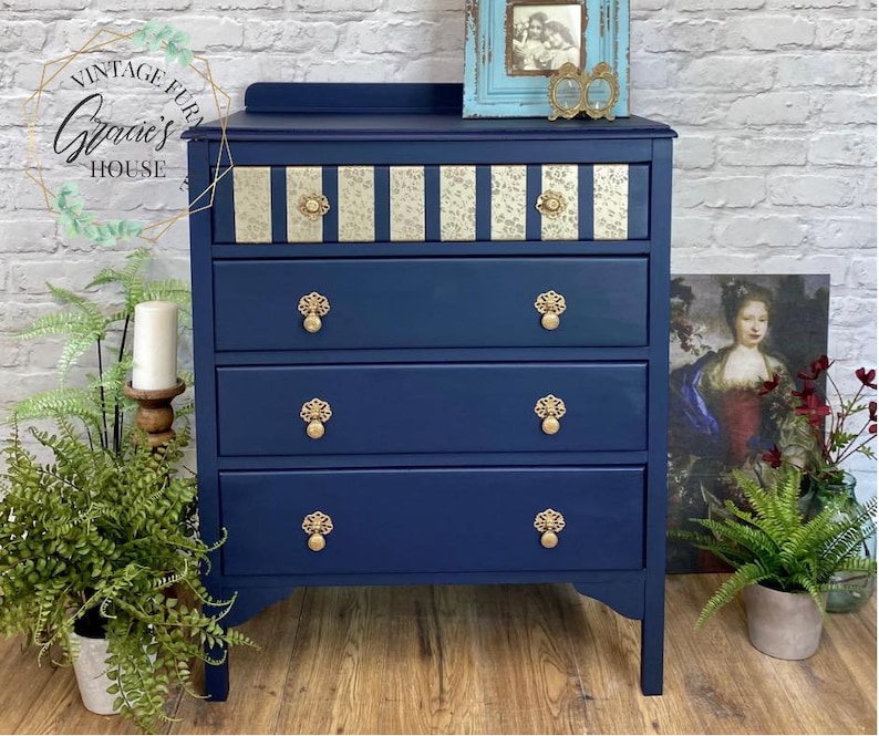 May include: A navy blue chest of drawers with four drawers and gold-toned hardware. The top drawer has a decorative gold and white patterned paper inlay. The chest of drawers is sitting on a wooden floor.