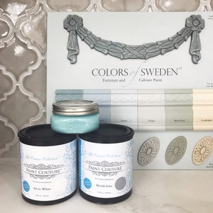 May include: Two paint cans, one labelled "Arctic White" and the other "British Grey", are displayed on a white surface. The cans are part of the "The Couture Collection!" line of Paint Couture. A glass jar of light blue paint is in the background, along with a colour chart for Colors of Sweden furniture and cabinet paint.