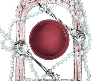 May include: Two silver spoons with ornate handles and a seashell design rest on a red lidded jar. The jar and spoons are surrounded by a pearl necklace and a pink ornate frame.