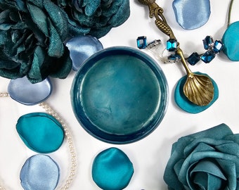 May include: A teal blue glass jar with a metallic sheen, surrounded by teal blue and light blue fabric flower petals, a gold spoon with a shell design, and a necklace with blue gemstones.
