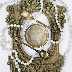 May include: A gold-colored metal bowl with a swirling pattern sits inside a decorative, ornate frame. A pearl necklace surrounds the frame, and two gold-colored spoons are placed on top of the frame.