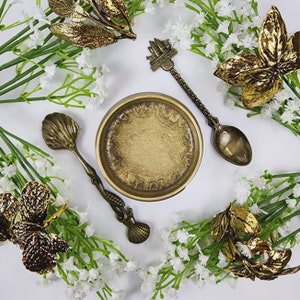 May include: A gold-toned metal bowl with a swirling pattern sits on a white surface surrounded by sprigs of white flowers and gold-toned leaves.