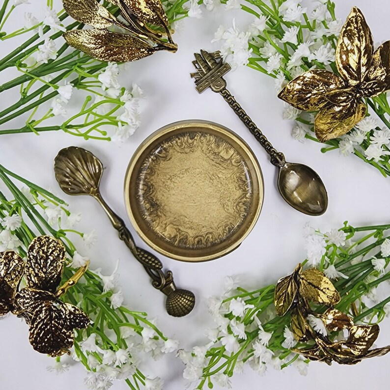 May include: A gold-toned metal bowl with a swirling pattern sits on a white surface surrounded by sprigs of white flowers and gold-toned leaves.