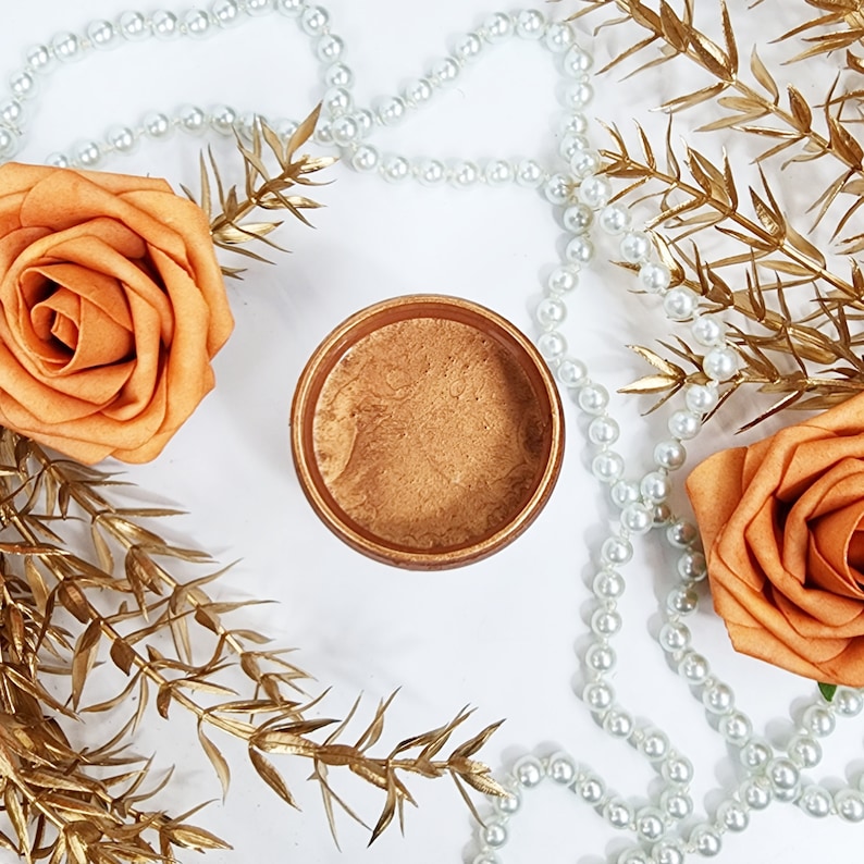 May include: A small round container of metallic gold paint with a textured surface. The container is surrounded by white pearl beads and gold-colored branches.
