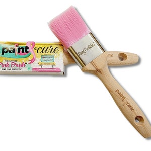 May include: A pink paint brush with a wooden handle and a silver ferrule. The brush is labelled "Paint Couture!" and "paint cure". The packaging for the brush is also visible and says "The Painters "Pink Brush" 2" Flat Fine Synthetic".