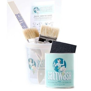 May include: A white can of Saltwash paint with a blue label featuring a woman in a blue dress and a white hat. The label also includes the text "Saltwash" and "For a layered sun & salt air soaked look". The can is surrounded by paintbrushes, a sanding sponge, and a white plastic container.