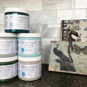 May include: Five jars of paint in various shades of green, blue, and grey. The jars are labelled "The Couture Collection Paint Couture" and each jar has a different colour name. The jars are arranged in two rows on a countertop.