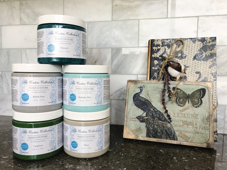 May include: Five jars of paint in various shades of green, blue, and grey. The jars are labelled "The Couture Collection Paint Couture" and each jar has a different colour name. The jars are arranged in two rows on a countertop.
