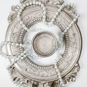 May include: A silver-colored ornate frame with a pearl necklace wrapped around it. A small silver-colored bowl sits in the center of the frame.