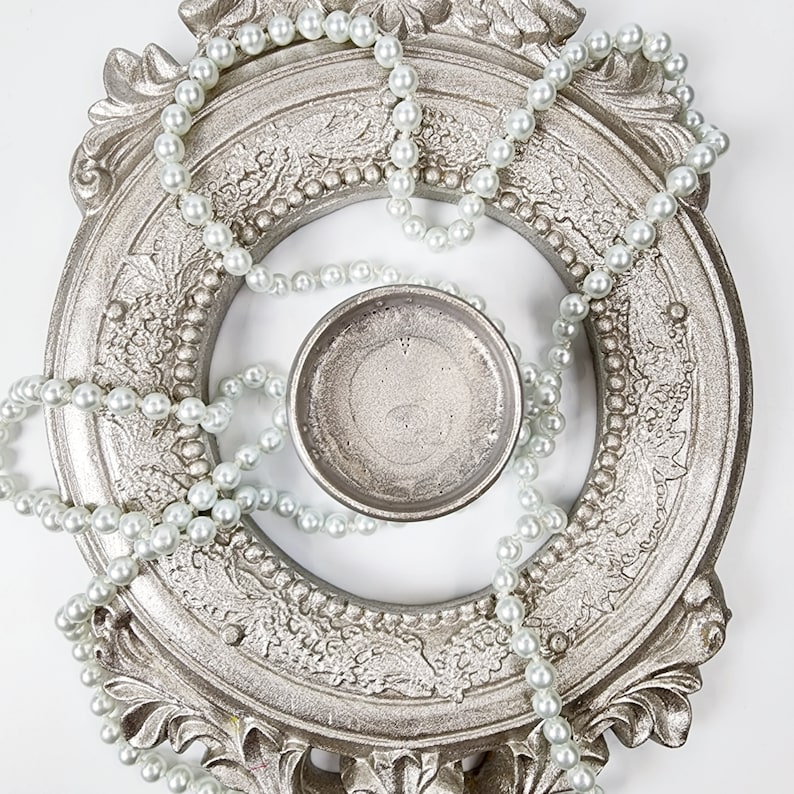 May include: A silver-colored ornate frame with a pearl necklace wrapped around it. A small silver-colored bowl sits in the center of the frame.