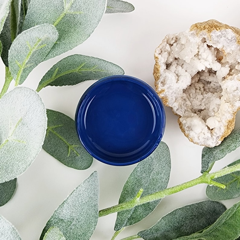 May include: A small, round, blue jar with a clear lid. The jar is sitting on a white surface with green leaves.
