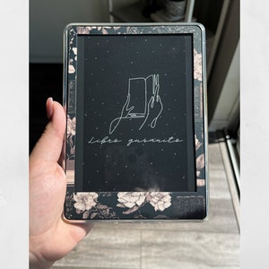 Op de afbeelding: Een zwarte e-reader met een bloemenontwerp op de rand. Het scherm toont een zwarte achtergrond met witte tekst die "libro gasarito" luidt en een lijntekening van een hand die een open boek vasthoudt.