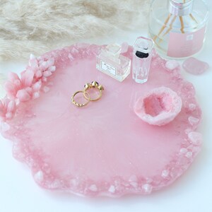 May include: A pink resin tray with a crystal-like border and accents. The tray holds gold rings, perfume bottles, and a geode. Decorative items are arranged on a white surface, creating a stylish display.