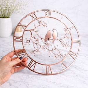 May include: Rose gold metal wall clock with Roman numerals and a decorative branch with two birds. The clock is round and has an open design, with the birds perched on a branch in the center.