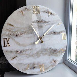Peut inclure: Une horloge murale ronde avec un cadran marbr&eacute; blanc et beige. Des chiffres romains dor&eacute;s indiquent les heures, et des aiguilles dor&eacute;es indiquent l'heure. Le cadran est orn&eacute; de petits cristaux et pierres scintillantes.