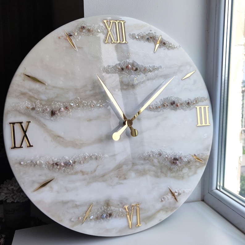 Peut inclure: Une horloge murale ronde avec un cadran marbr&eacute; blanc et beige. Des chiffres romains dor&eacute;s indiquent les heures, et des aiguilles dor&eacute;es indiquent l'heure. Le cadran est orn&eacute; de petits cristaux et pierres scintillantes.