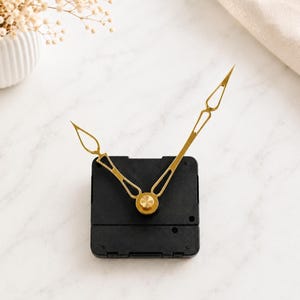 May include: A black square clock movement mechanism featuring two elongated, gold-toned metal clock hands with open, diamond-shaped tips. The hands are attached to a central gold-colored circular cap, positioned against a plain, light-colored surface.