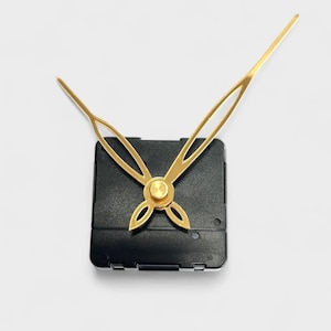 May include: A clock movement with a black square body and gold-colored hands. The hands are stylized with a decorative design. The hour hand is a stylized arrow shape, and the minute hand is a long, slender design.