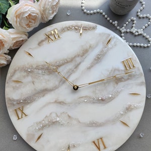 May include: A round, white wall clock with gold Roman numerals and hands. The clock face has a marbled, cream and beige design with embedded crystals. The clock is surrounded by decorative elements, including flowers and a candle.