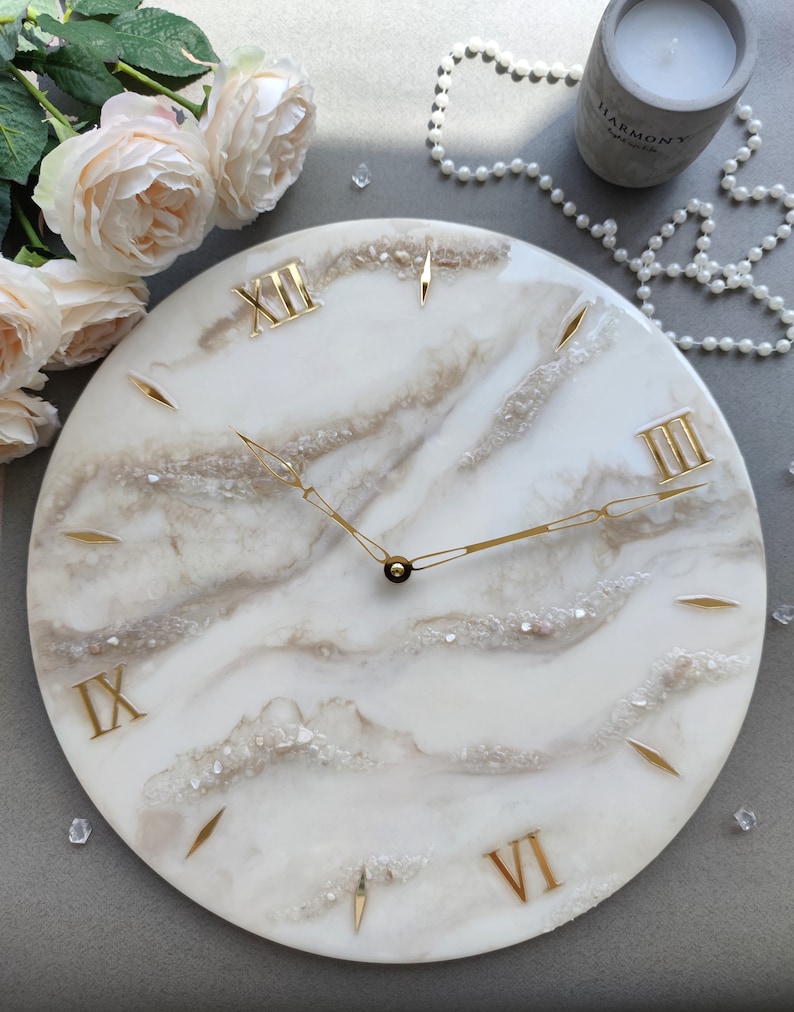 May include: A round, white wall clock with gold Roman numerals and hands. The clock face has a marbled, cream and beige design with embedded crystals. The clock is surrounded by decorative elements, including flowers and a candle.