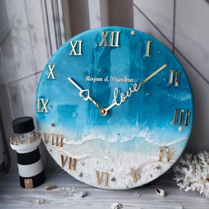May include: A round wall clock with a blue and white ocean-themed design. The clock features gold Roman numerals and hands, with the word "love" and the names "Rajan & Vandna" in gold script. A decorative lighthouse is visible.