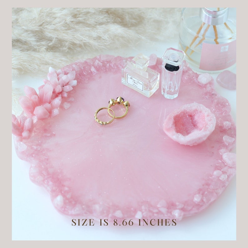 May include: A pink resin tray with a crystal-like border, holding two gold rings, perfume bottles, and a geode. The tray is 8.66 inches in diameter. The tray has a smooth surface with a glittery effect.