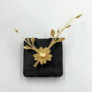 May include: A gold-coloured clock mechanism with a floral design. The clock face features a flower in the centre and leaf-like accents extending outwards. The clock's body is a black square.