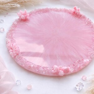 May include: A round, pink resin tray with a glossy finish. The tray is decorated with small, clear crystals around the edge and has several small, pink floral accents. The tray is approximately 10 inches in diameter.