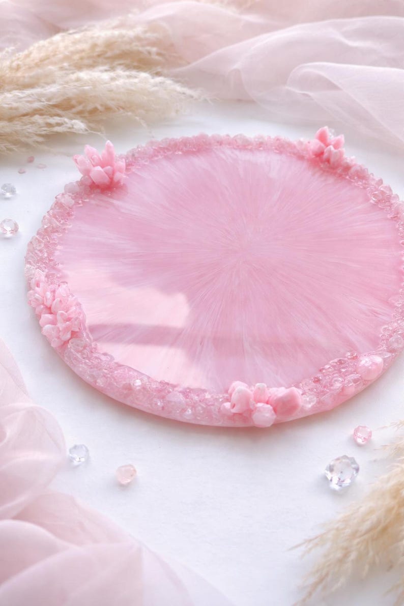 May include: A round, pink resin tray with a glossy finish. The tray is decorated with small, clear crystals around the edge and has several small, pink floral accents. The tray is approximately 10 inches in diameter.