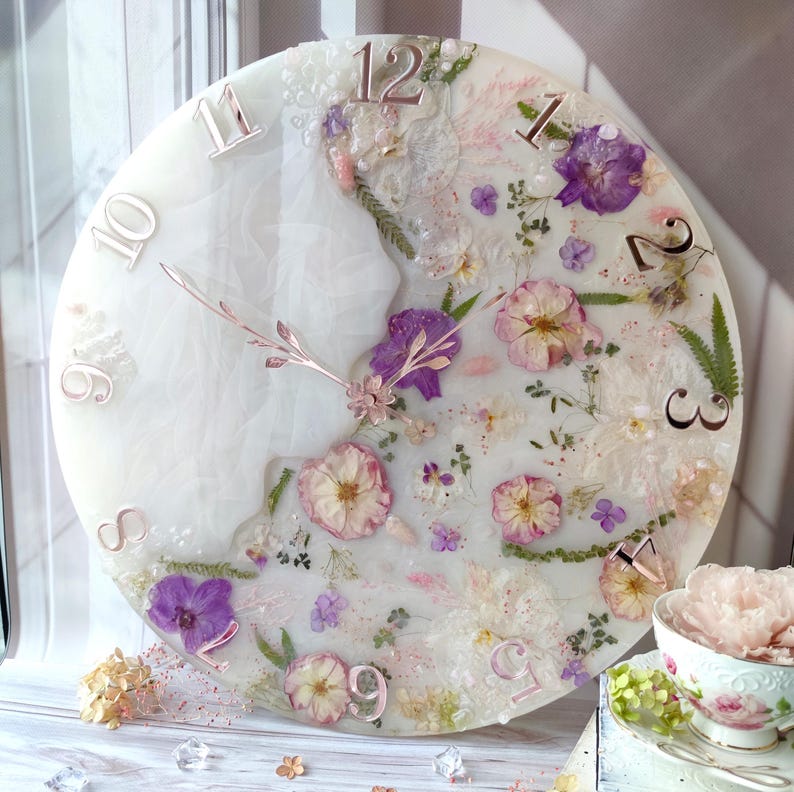 May include: A round, white clock with embedded dried flowers in shades of purple, pink, and white. The clock face features rose gold numerals and hands. The clock is a decorative item.