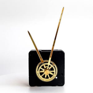 Puede incluir: Un mecanismo de reloj negro con manecillas y un engranaje dorados. Las manecillas son largas y delgadas, y el engranaje es circular con dientes intrincados. El mecanismo del reloj tiene forma cuadrada.