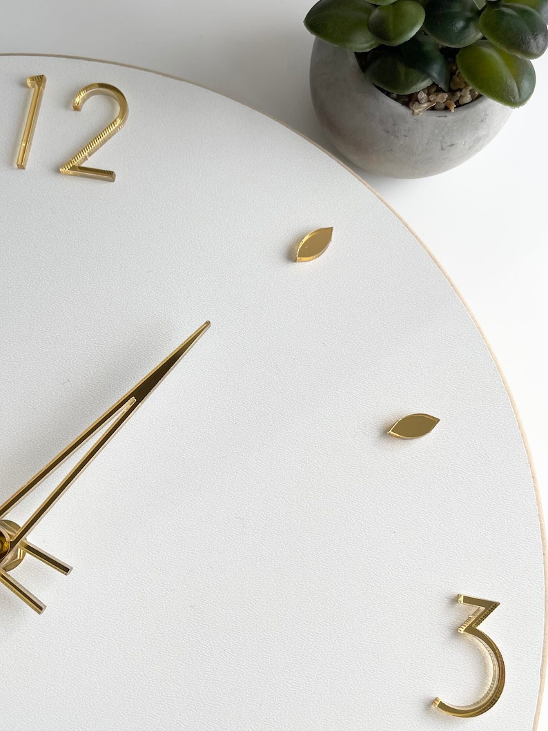 Peut inclure: Gros plan sur une horloge murale blanche avec des aiguilles et des chiffres dor&eacute;s. Le cadran de l'horloge a une surface textur&eacute;e. Les chiffres 12 et 3 sont visibles, ainsi que des marqueurs dor&eacute;s en forme de feuille. Une petite plante en pot est en arri&egrave;re-plan.