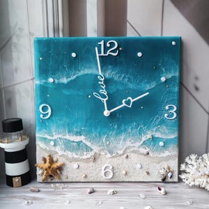 Peut inclure: Horloge murale carr&eacute;e avec un motif de sc&egrave;ne de plage. Le cadran de l'horloge repr&eacute;sente un oc&eacute;an turquoise avec des vagues blanches, une plage de sable et des chiffres blancs. Le mot "love" est &eacute;crit en cursive sur le cadran. Une &eacute;toile de mer et des coquillages sont sur la plage.