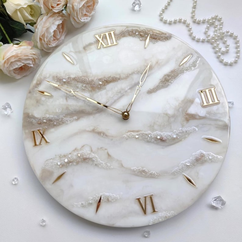 May include: A round wall clock with a white and beige marbled design. The clock features gold Roman numerals, gold hands, and gold markers. The clock is surrounded by decorative elements, including flowers and crystals.