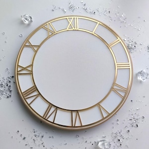 May include: A circular clock face with gold Roman numerals and a gold border, set against a white background. The clock face is decorated with clear, crystal-like embellishments. The clock face is a decorative item.