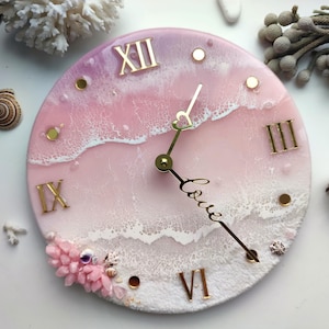 Könnte beinhalten: Eine runde Wanduhr mit einem rosa-weißen Harz-Ozeanwellen-Design. Das Zifferblatt enthält goldene römische Ziffern an den Positionen zwölf, drei, sechs und neun sowie goldene Zeiger, einer in Herzform und einer mit dem Wort love.
