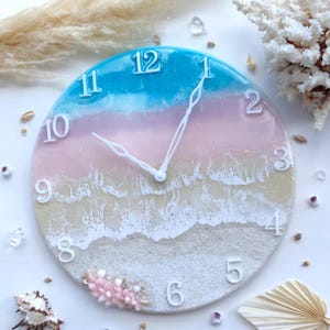 May include: A round wall clock featuring a resin art design that mimics a beach scene with layers of turquoise blue, soft pink, and sandy beige. The clock face includes white raised numerals and white clock hands, with small pink crystals.
