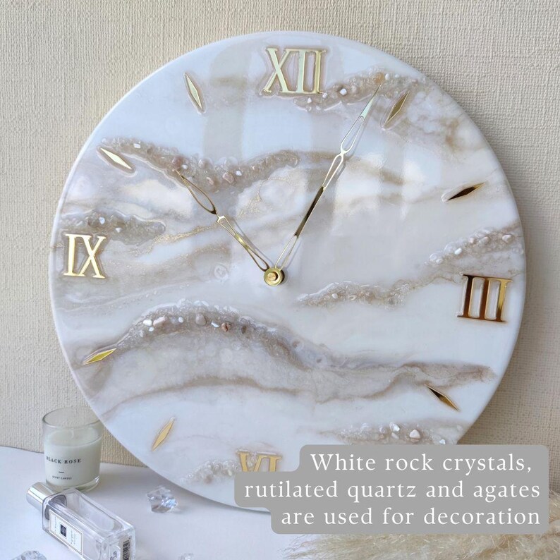 May include: Round white wall clock with gold Roman numerals and hands. The clock face features white rock crystals, rutilated quartz, and agates. The text on the image says: White rock crystals, rutilated quartz and agates are used for decoration.