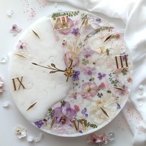May include: A round wall clock with a white background, featuring embedded dried flowers in shades of pink, purple, and white. The clock has gold Roman numerals and gold hands. The clock measures approximately 12 inches in diameter.